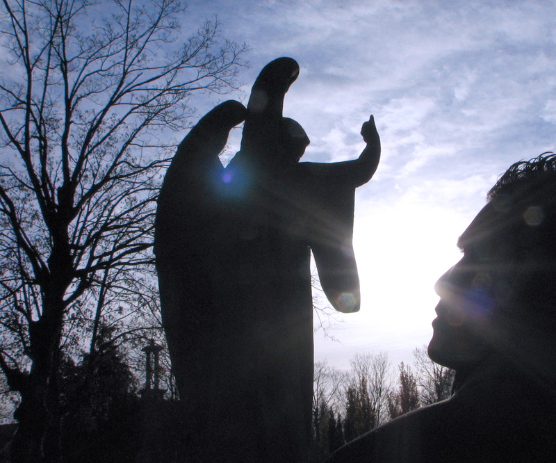 Silhouettes of a man and an angel's statue, against the light of the sun.